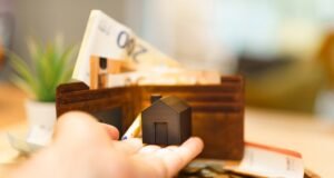 Šta sve stoji iza cene stambenog kredita i kako prepoznati skrivene troškove? Model kuće u ruci pored novčanika, novčanica evra i kovanica