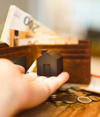 Šta sve stoji iza cene stambenog kredita i kako prepoznati skrivene troškove? Model kuće u ruci pored novčanika, novčanica evra i kovanica
